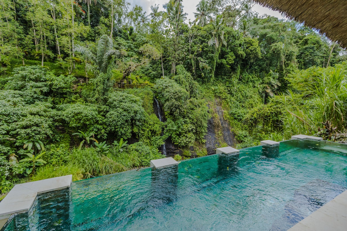 An infinity glass pool melds seamlessly with the vibrant greenery surrounding it. Lush jungle vegetation and cascading waterfalls are visible in the background, creating a serene ambience in this secluded hideaway.