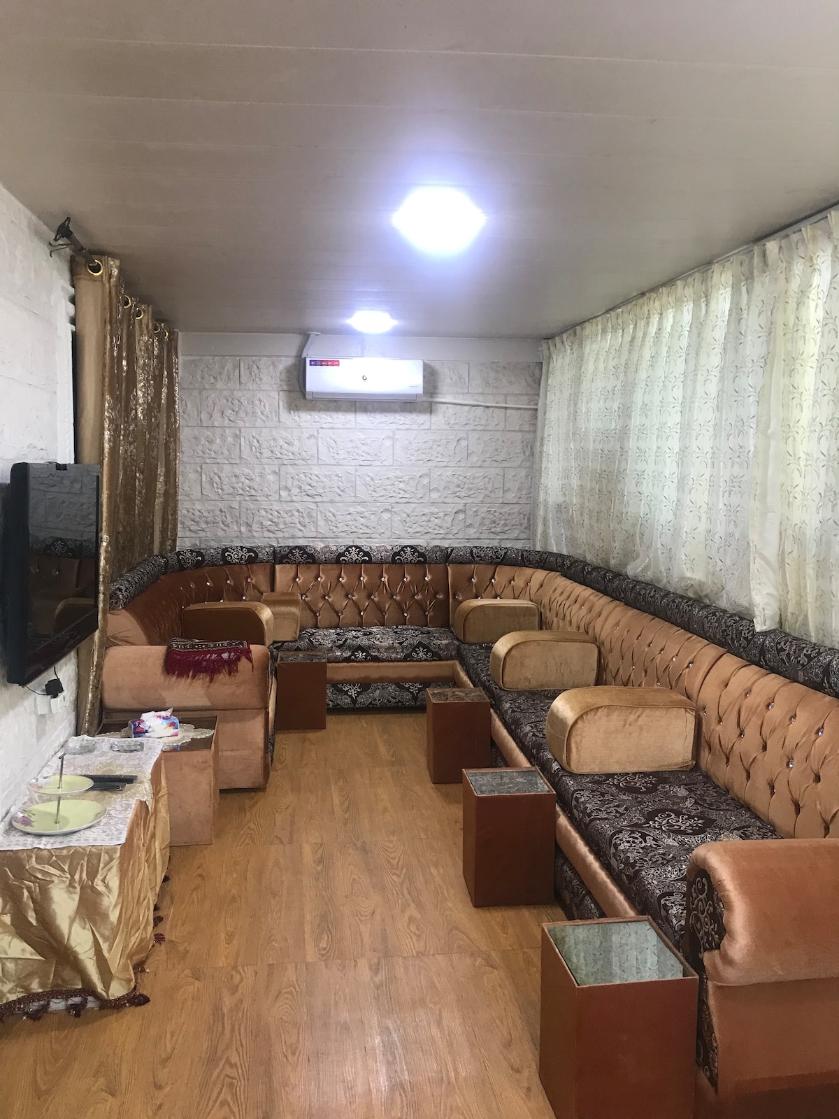 A cozy living area is arranged with L-shaped seating upholstered in warm tones. Small glass tables complement the seating, and light streams through sheer curtains, enhancing the space. A television is mounted on the wall, providing a focal point for relaxation.