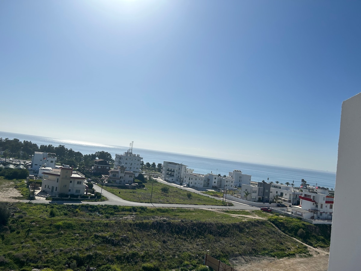 A panoramic view showcases the coastline, featuring golden sands and the shimmering blue sea under a clear sky. Buildings are visible in the foreground, surrounded by greenery and open spaces, presenting a serene atmosphere for relaxation.