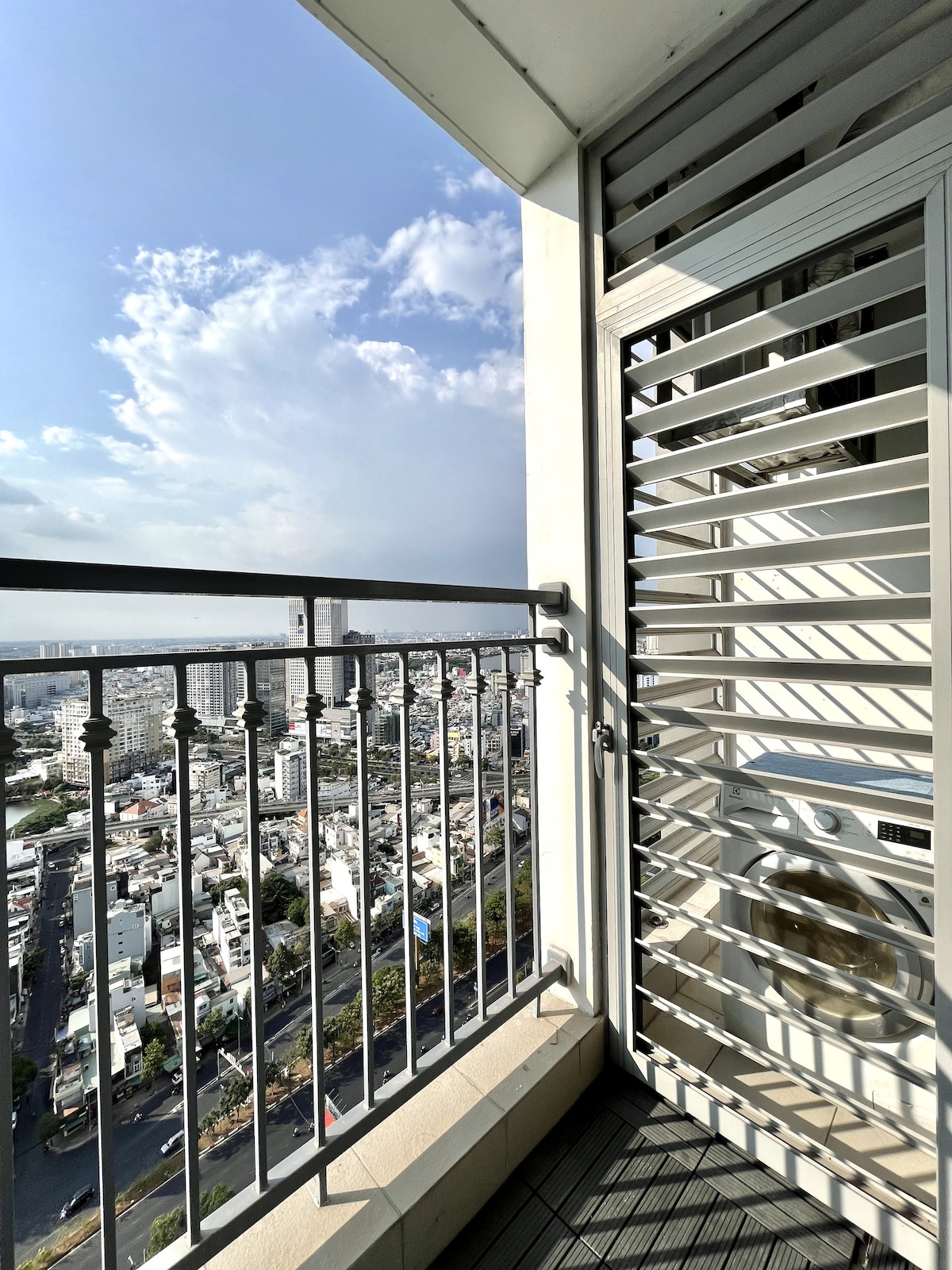A balcony is shown with a metal railing that offers a view of the city below. The corner of a washing machine is visible, along with slatted panels that provide ventilation and light. Bright sunlight casts shadows on the balcony floor.