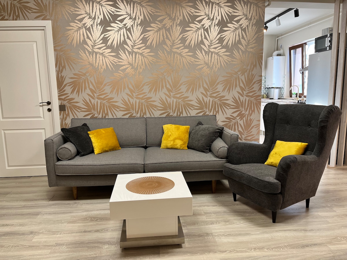 A modern living space features a gray sofa adorned with yellow and black cushions, complemented by a dark gray armchair. A white coffee table with a circular wooden centerpiece sits in front of the seating area. The walls are decorated with a subtle gold leaf pattern.