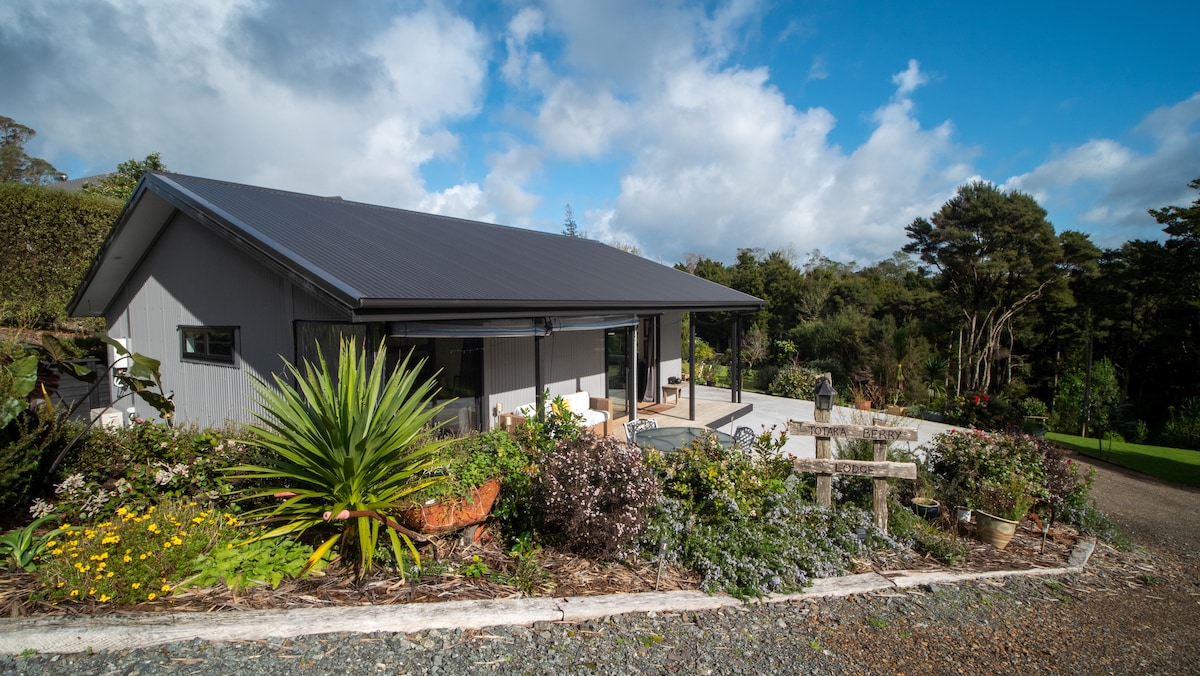 The exterior of the lodge is presented, showcasing a single-story building framed by lush native bush and vibrant flowerbeds. A spacious deck is visible, providing access to outdoor seating, while the surrounding landscape offers a peaceful blend of greenery and cultivated gardens.