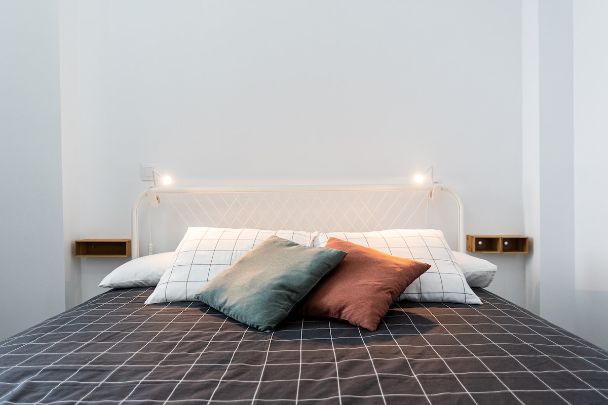 A modern bedroom features a spacious bed dressed in a dark grid-patterned duvet. Soft pillows in varying shades rest at the head of the bed, with bedside lights mounted on the walls providing a gentle glow.
