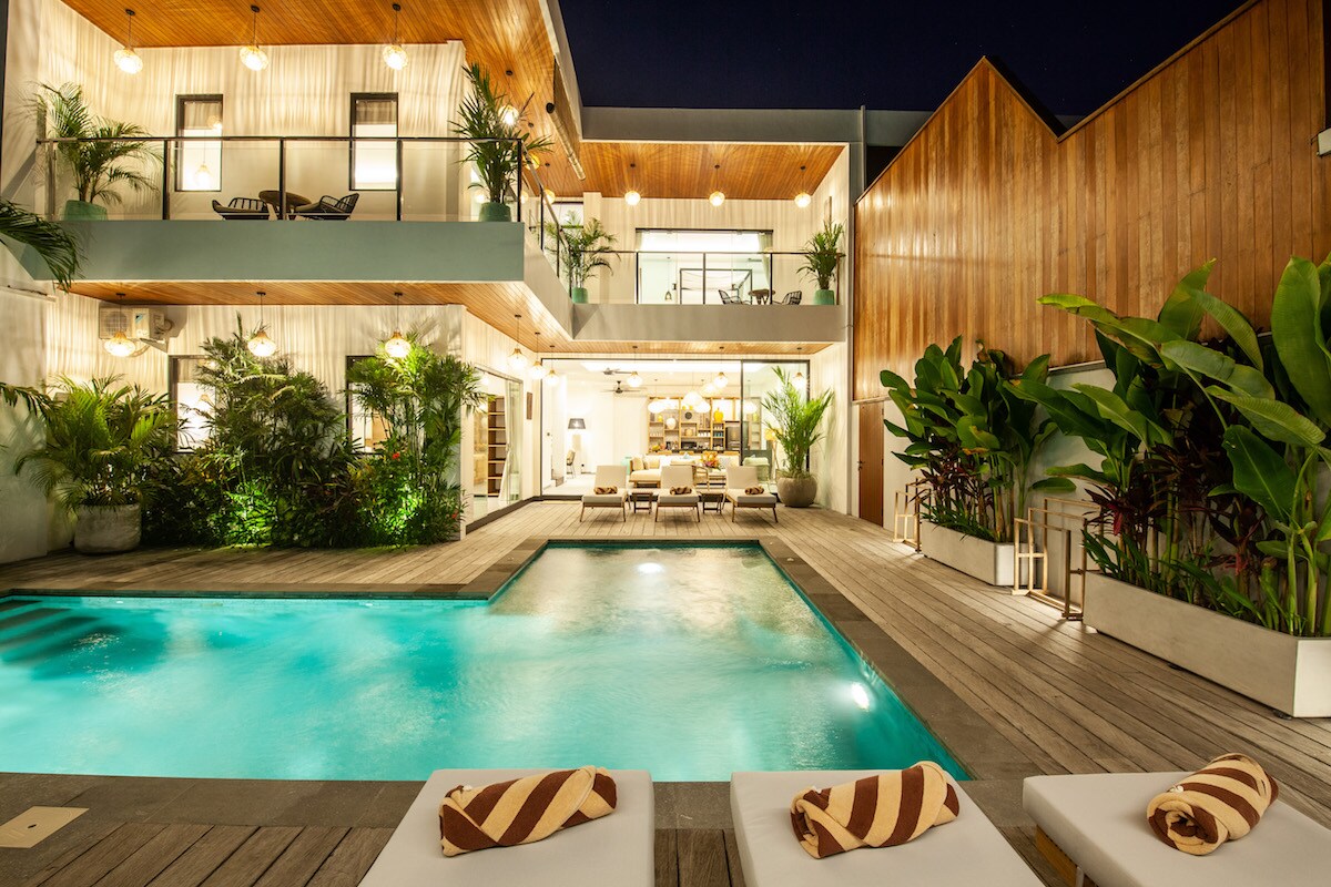 The exterior of the villa is illuminated by soft lighting, showcasing a private pool with surrounding wooden decking and sunbeds. Lush tropical plants frame the image, while the two-story structure features large windows, providing a glimpse into the spacious living area and outdoor dining space.