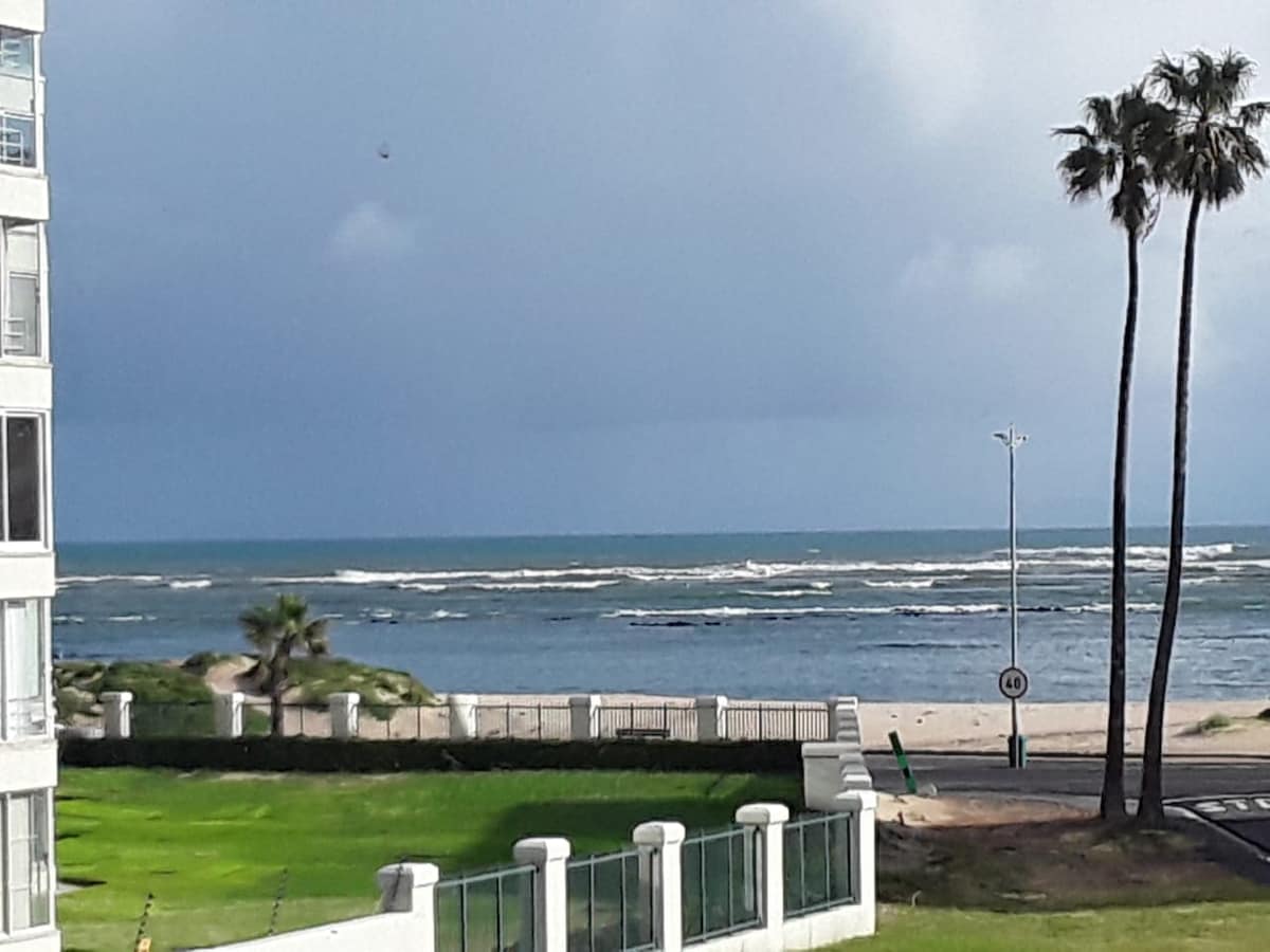 A view of the ocean showcases rolling waves and a sandy beach, framed by two palm trees in the foreground. The grassy area leads to the beach path, visible in the lower part of the image, with a gentle coastal landscape in the distance.