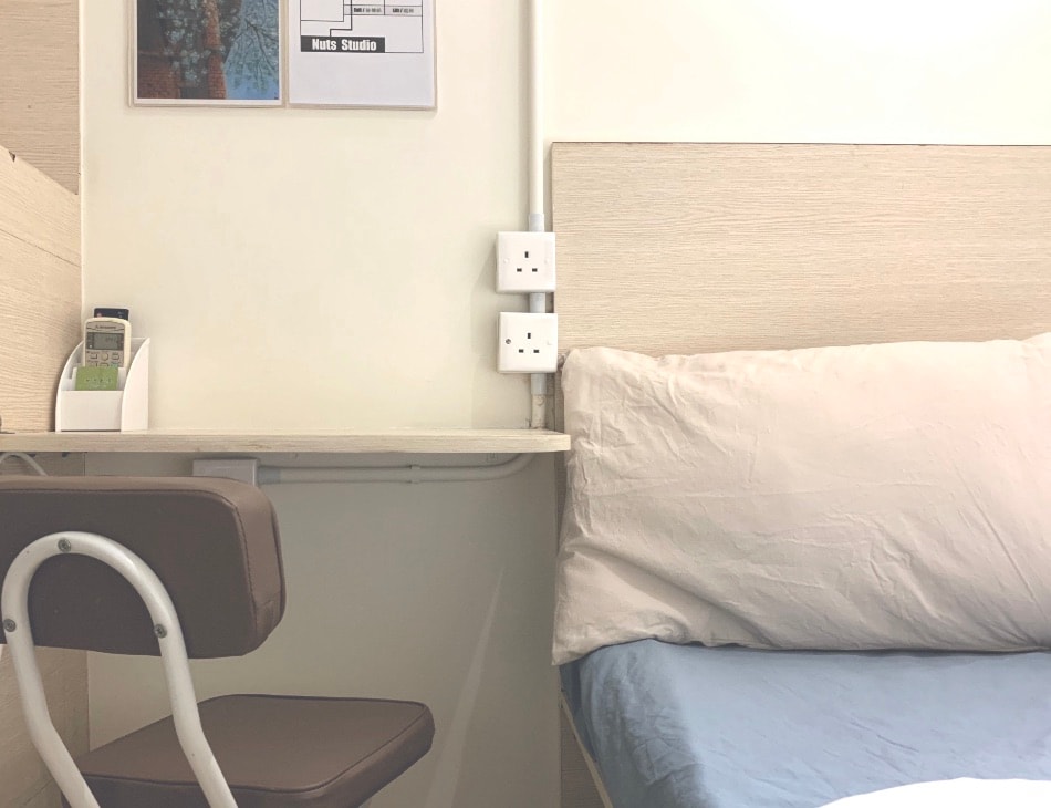 A compact corner of the studio features a spacious bed with a neatly arranged light-colored pillow. A small desk with a chair is positioned nearby, alongside electrical outlets for convenience. A clock and informational chart are visible on the wall above the desk.