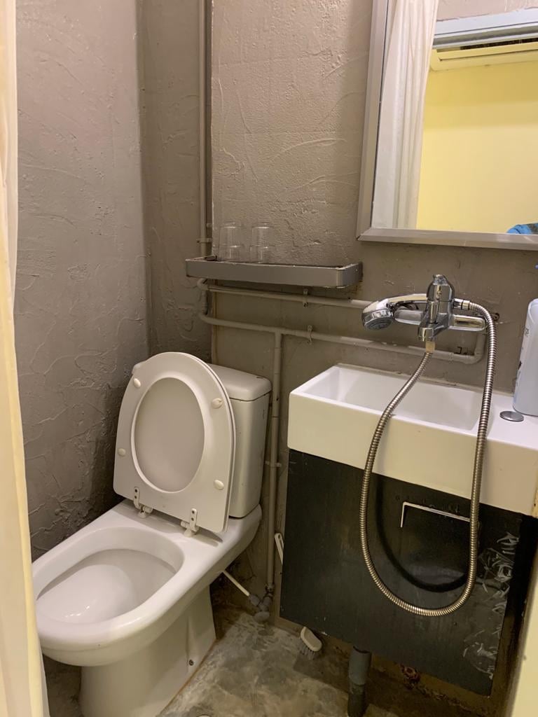 The image displays a compact bathroom featuring a toilet alongside a small sink with a modern faucet. A mirror hangs above the sink, and a shelf can be seen above the toilet holding two glass tumblers. The walls are finished in a light grey texture.