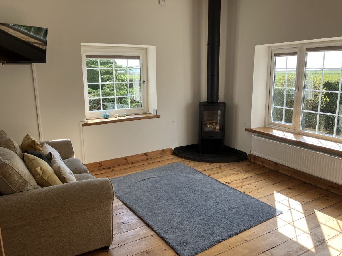 A large open sitting room is highlighted by natural light streaming through two windows with lush greenery visible outside. A wood burner stands in the corner beside a comfortable sofa, and a soft area rug is placed on the wooden floor.