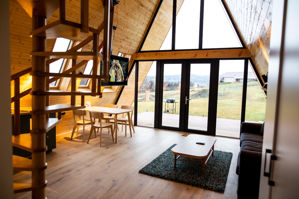 An A-frame structure features a spacious living area with large windows allowing natural light. A spiral staircase leads to an upper level. A cozy couch, wooden dining table with chairs, and a small coffee table are present, complementing the warm wooden interior.