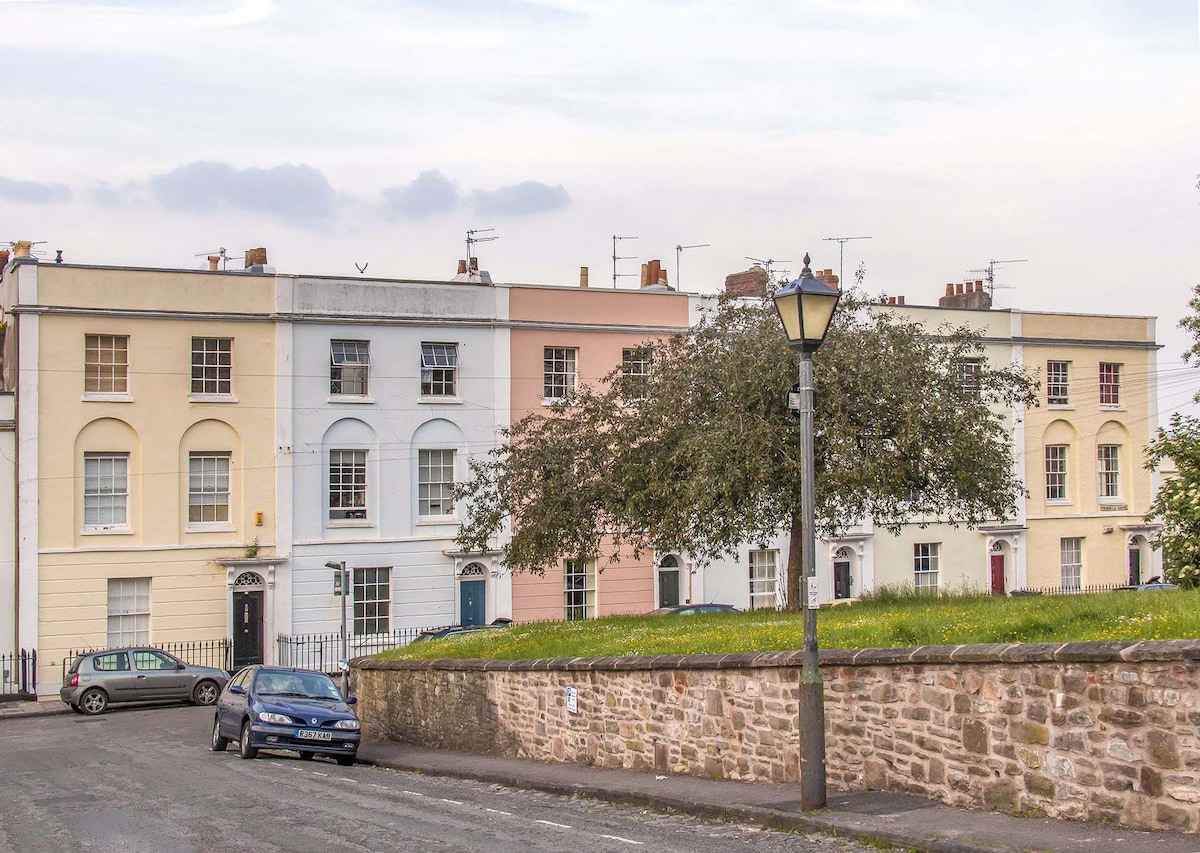 The image showcases a row of late Georgian and early Victorian townhouses in pastel shades. Each building features distinct architectural details, including large windows and ornate doors. A grassy area is visible in the foreground, with a street lamp providing soft illumination.