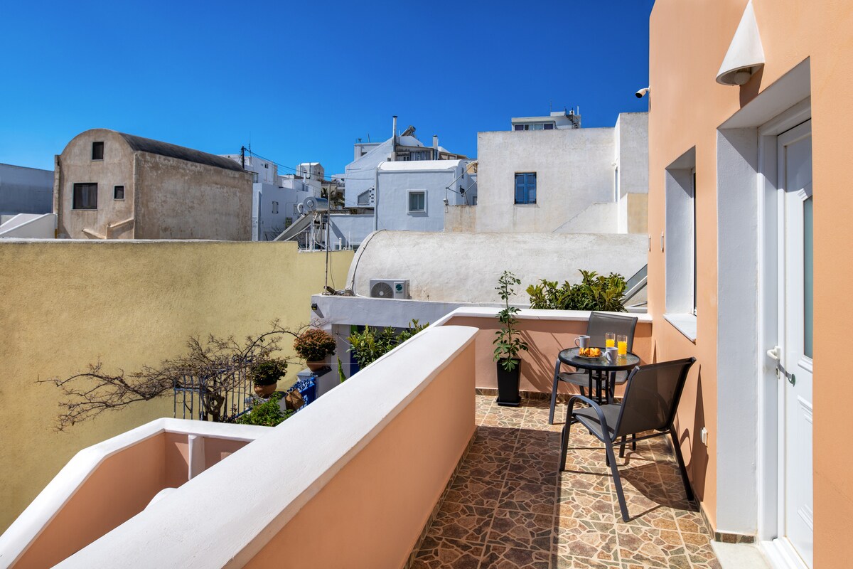 A private terrace is visible, featuring a small table with two chairs and a view of surrounding buildings. Potted plants add greenery to the space, while the textured floor adds visual interest. Bright blue skies complement the warm tones of the adjoining structures.