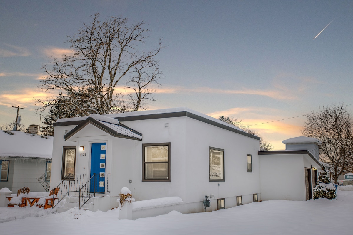 A modern, single-story home with a white exterior is shown bathed in soft sunset light. Snow covers the ground, while a blue front door and large windows add contrast. A bare tree in the background frames the serene winter scene.
