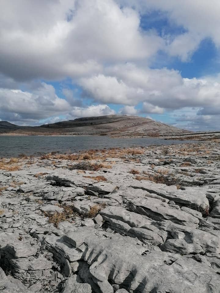 A rugged karst landscape is depicted, featuring sprawling limestone formations and patches of dried grass. In the background, a gentle hill rises under a partly cloudy sky, with varying shades of blue peeking through the clouds, creating a serene natural setting.