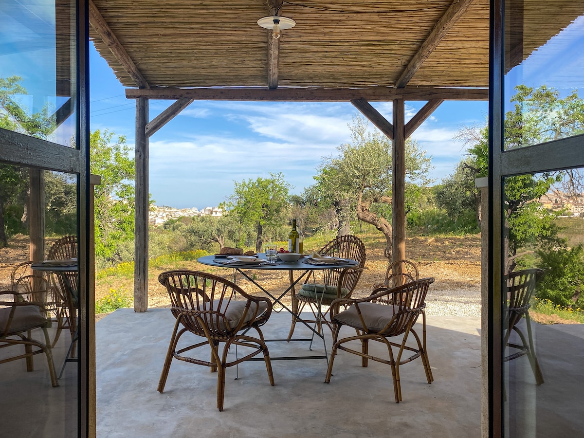 A shaded outdoor terrace features a dining table surrounded by rattan chairs, providing a space for meals with views of the surrounding countryside. Sunlight filters through the wooden structure, highlighting the natural scenery and olive trees beyond.