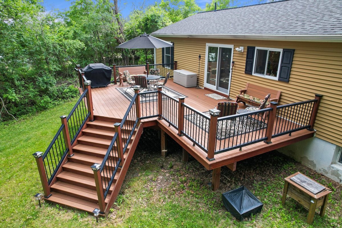A spacious back deck is displayed, featuring a seating area with multiple chairs and a table. A gas grill is positioned nearby, alongside an umbrella for shade. The deck is surrounded by greenery, providing a natural setting for outdoor relaxation.