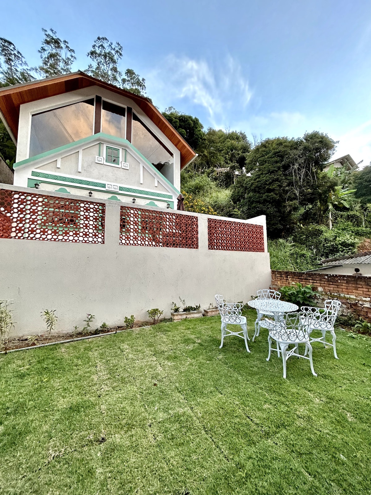 The exterior of the house is shown, featuring a modern design with large windows and a sloped roof. A manicured lawn surrounds a white patio set with four chairs and a round table. Lush greenery is visible in the background, complementing the serene environment.