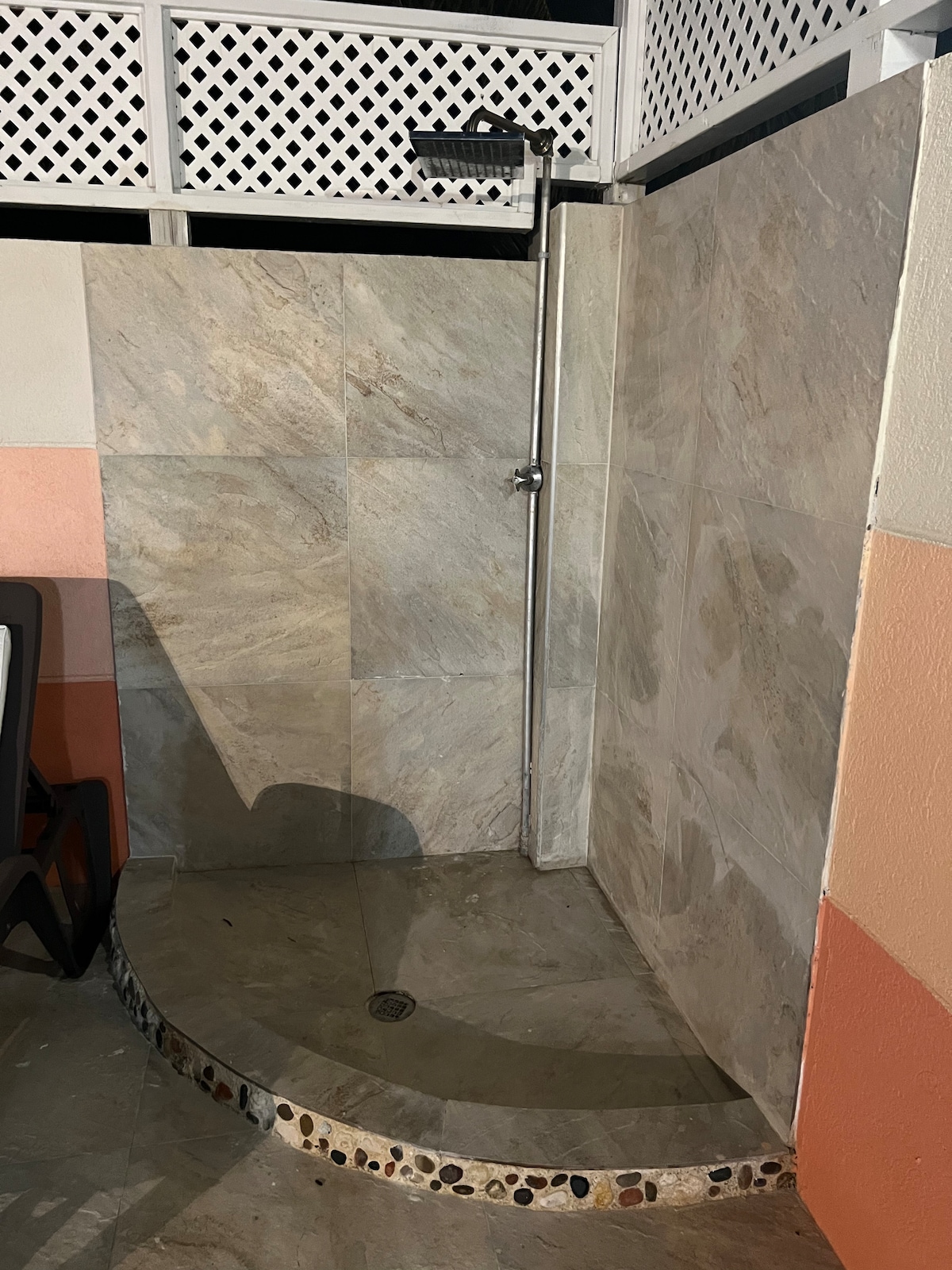 An outdoor shower area is depicted, featuring smooth stone walls and a circular shower basin. A wall-mounted showerhead is shown above, and the ground is tiled with a combination of warm and cool tones, enhancing the serene ambiance.