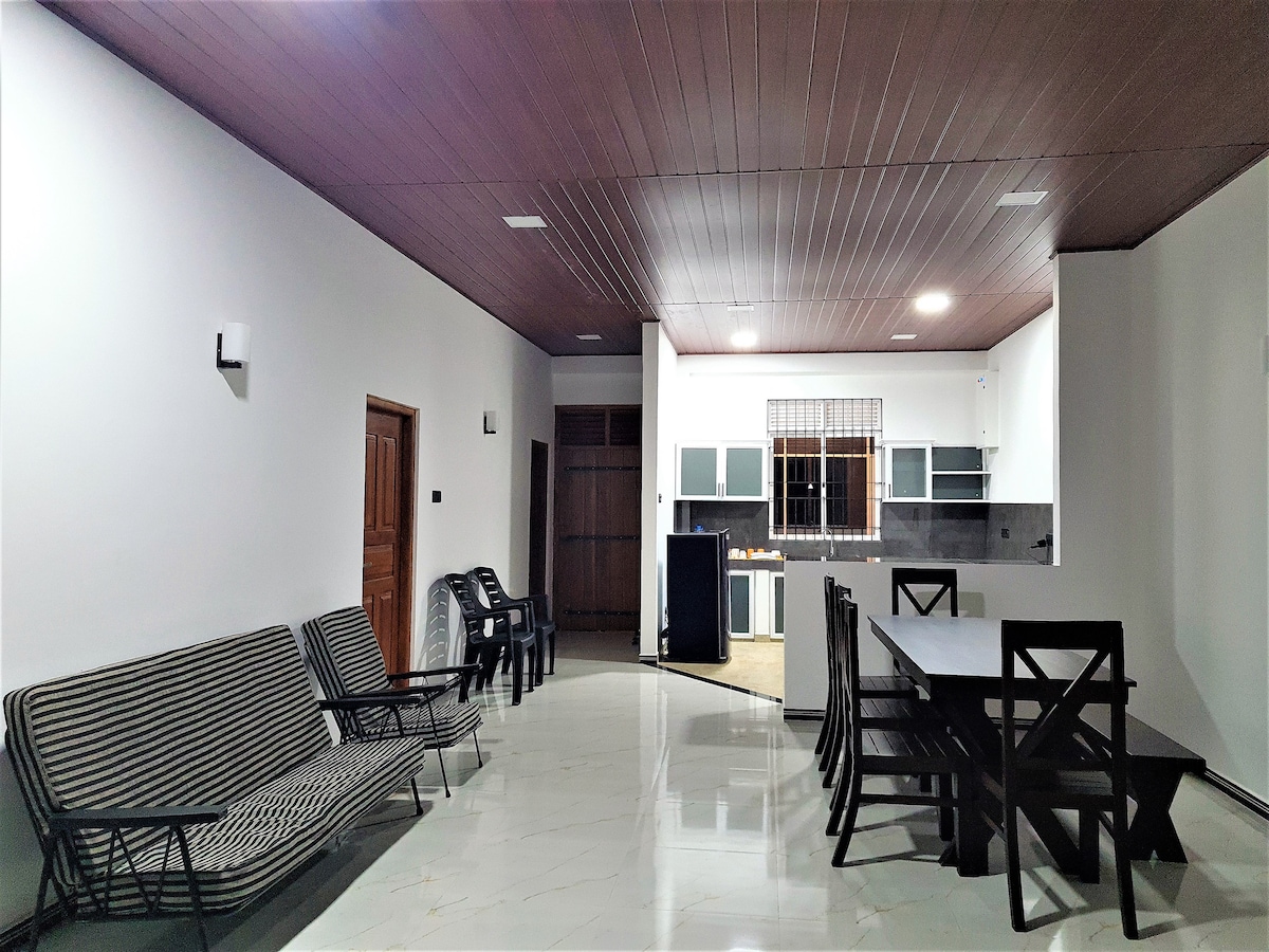A spacious living area features a striped sofa and several black plastic chairs. A long wooden dining table is surrounded by matching chairs. The open kitchen area is visible in the background, showcasing cabinets and modern appliances against a neutral wall.