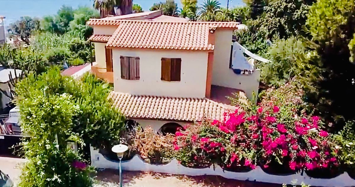 An aerial view captures the villa surrounded by lush greenery and vibrant pink bougainvillea. The two-story structure features a tiled roof and wooden shutters, with shaded areas offering a sense of privacy. The tranquil setting near the beach is highlighted by the lush garden.