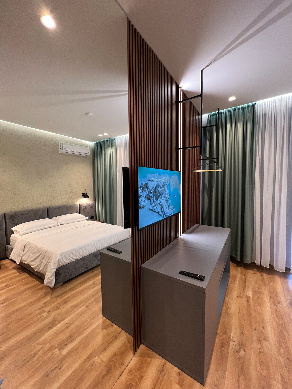 The room features a cozy sleeping area with a double bed, separated from the living space by a wooden partition. A wall-mounted television is positioned on the partition, alongside a sleek console. Soft, floor-length curtains frame the window, allowing for a gentle flow of natural light.