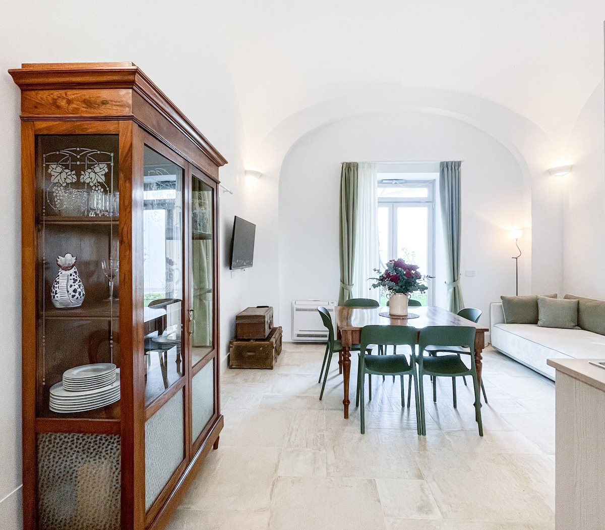 A spacious living area is showcased, featuring a wooden cabinet displaying dishes and decorative items. A dining table surrounded by green chairs is positioned beside a cozy seating area. Natural light enters through large windows, illuminated by soft lighting fixtures.