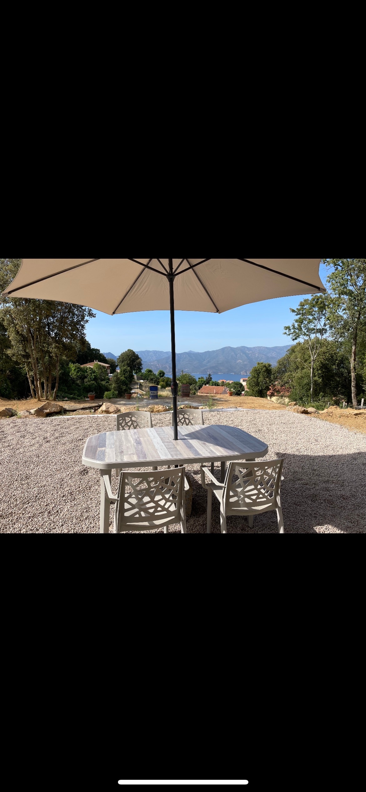 An outdoor dining area is set under a large umbrella, featuring a long table surrounded by several chairs. A scenic view of distant mountains and lush greenery is visible in the background, creating a serene environment.