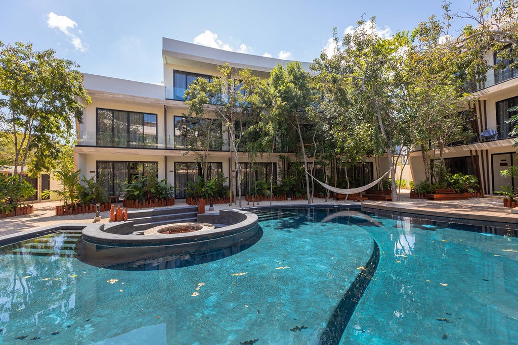 A serene pool area features a natural stone fire pit surrounded by clear water. The complex's architecture showcases large glass windows and tropical greenery. A hammock is seen between two trees, providing a tranquil space for relaxation.