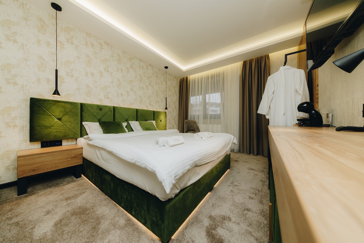 A spacious bedroom features a plush green upholstered bed with white linens and decorative pillows. Soft lighting illuminates the room, which is accented by curtains and a warm-colored wall. A wooden dresser, a coffee maker, and a white robe are also present.