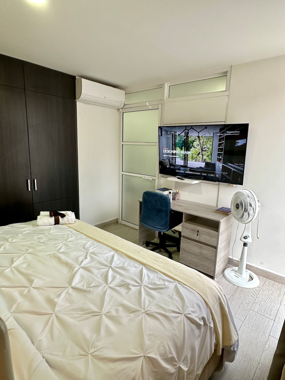 A neatly arranged room is equipped with a spacious bed covered in a light-colored quilt. A television is mounted on the wall, and a small desk with a chair is positioned adjacent to it. An air conditioning unit and a fan are visible, providing comfort.