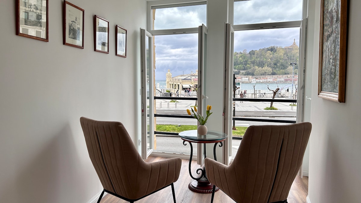 Two armchairs are positioned around a small round table, creating a cozy seating area. Large windows open to the scenic view of the waterfront, allowing natural light to fill the space. Decorative elements include framed photographs on the walls and a vase with fresh flowers.