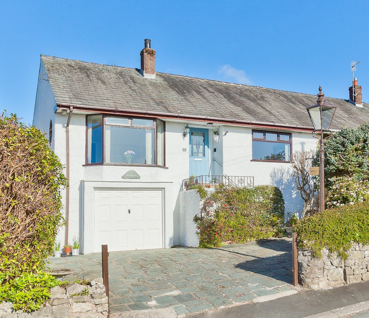 A light-colored bungalow is presented with a welcoming blue front door. The stone-paved driveway leads to a single garage below a well-maintained front garden, lined with greenery. Clear blue skies create a bright atmosphere around the property.