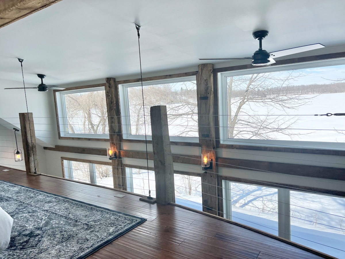 Large windows are framed by wooden beams, allowing for expansive views of the snowy landscape outside. Ceiling fans add a modern touch to the loft area, complemented by stylish light fixtures that provide soft illumination.