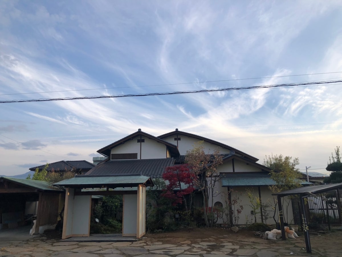 A traditional house is set against a backdrop of a clear sky with wispy clouds. The structure features a multi-tiered roof and a combination of light and dark exterior hues. Trees and shrubbery surround the property, creating a natural and serene atmosphere.