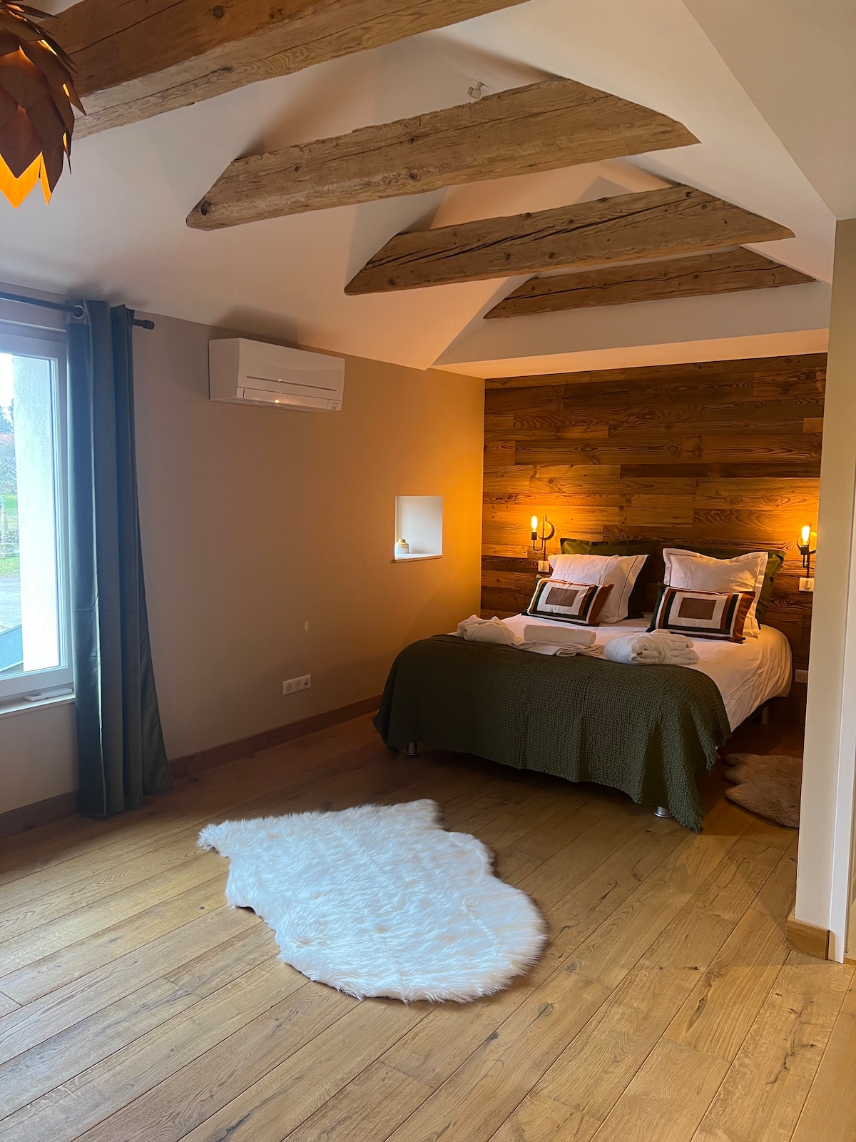 A cozy bedroom is highlighted by wooden beams overhead and warm wood accents on the wall. The spacious bed is adorned with neutral bedding and placed on a wooden floor, complemented by a soft area rug. Natural light enters from a large window.