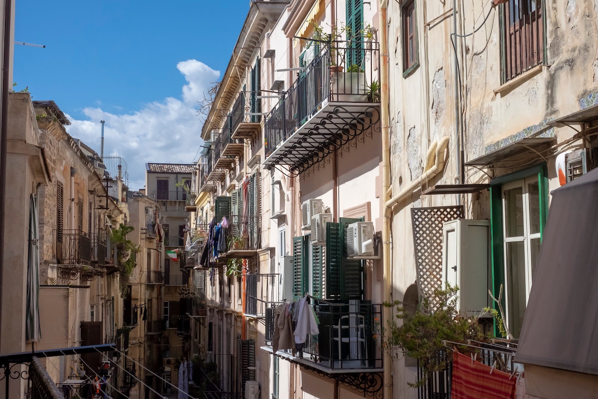 A narrow street in a historic neighborhood is lined with traditional buildings featuring balconies adorned with plants and laundry. Natural light illuminates the façades, showcasing varying architectural details and colors. Blue skies are visible above, contributing to the vibrant atmosphere of the area.