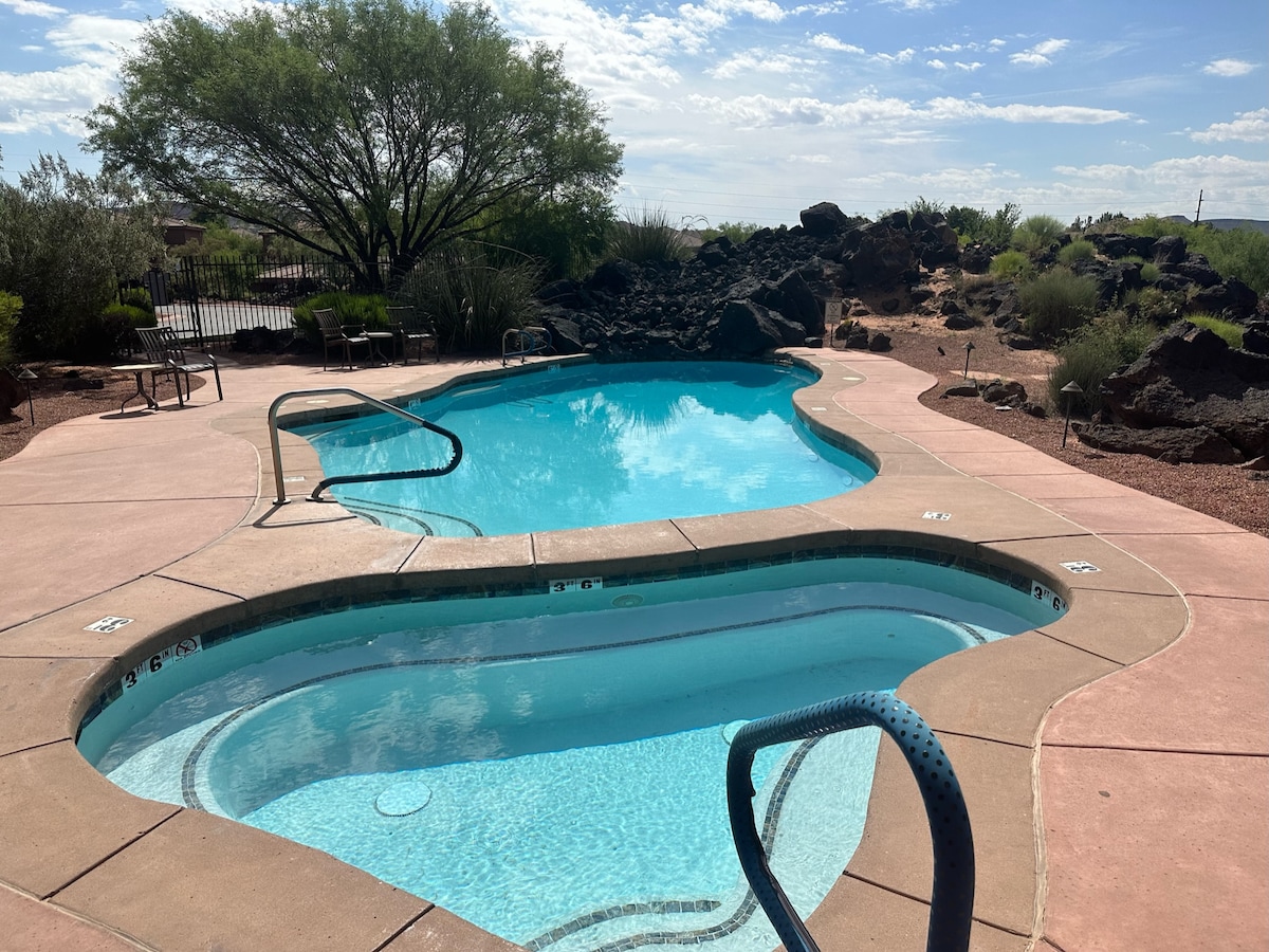 A community pool area features a uniquely shaped swimming pool alongside a hot tub. Smooth concrete edges surround the water, and lounge chairs are positioned nearby. Desert landscaping with low-lying vegetation and natural rock formations provide a scenic backdrop under a clear sky.