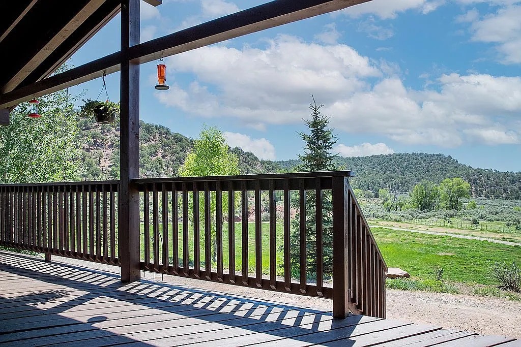 The view from a wooden deck showcases expansive greenery and rolling hills. A railing frames the scene, while the sky is partly cloudy with soft hues. Hummingbird feeders hang nearby, adding a touch of interest to the natural landscape.