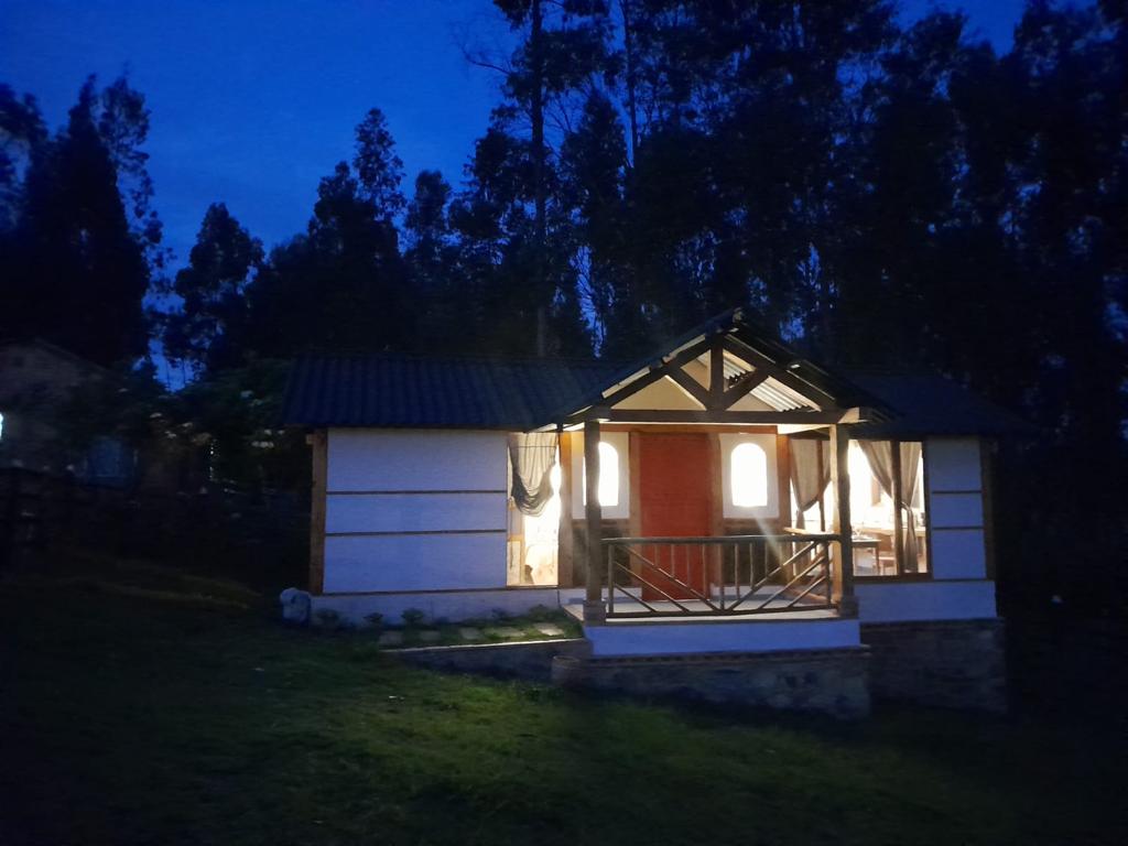 A cozy cabin is viewed at dusk, with warm light glowing from inside. The structure features a sloped roof and a welcoming front porch with a railing. Surrounding trees create a natural backdrop, enhancing the sense of tranquility and connection to nature.