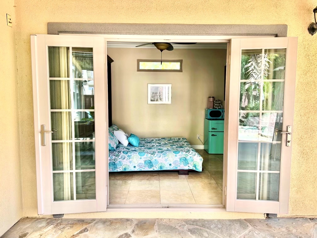 Double French doors open to reveal a bright interior space featuring a queen bed decorated with a floral-patterned comforter. A small turquoise mini fridge is visible in the corner, with soft lighting illuminating the room, and a piece of wall art is hung above the bed.