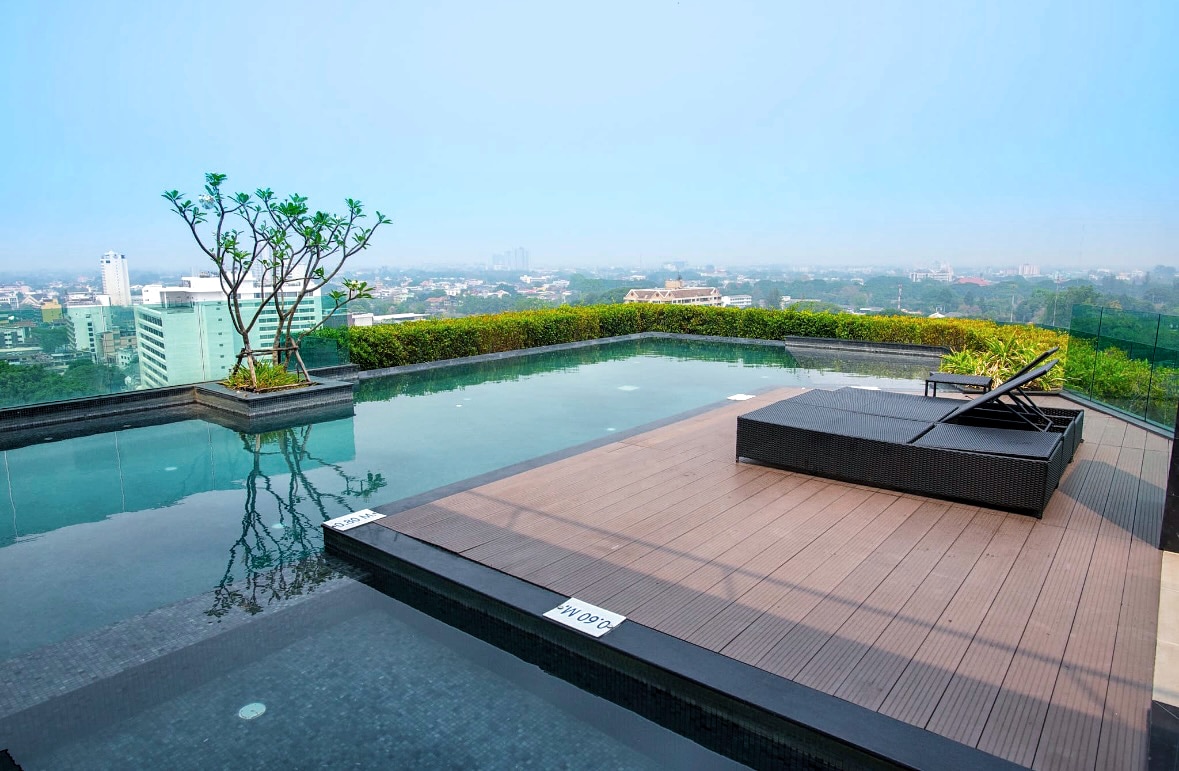 An infinity pool is featured on the rooftop, surrounded by lush greenery with city views in the background. A sun lounger is positioned on the wooden deck, inviting relaxation.