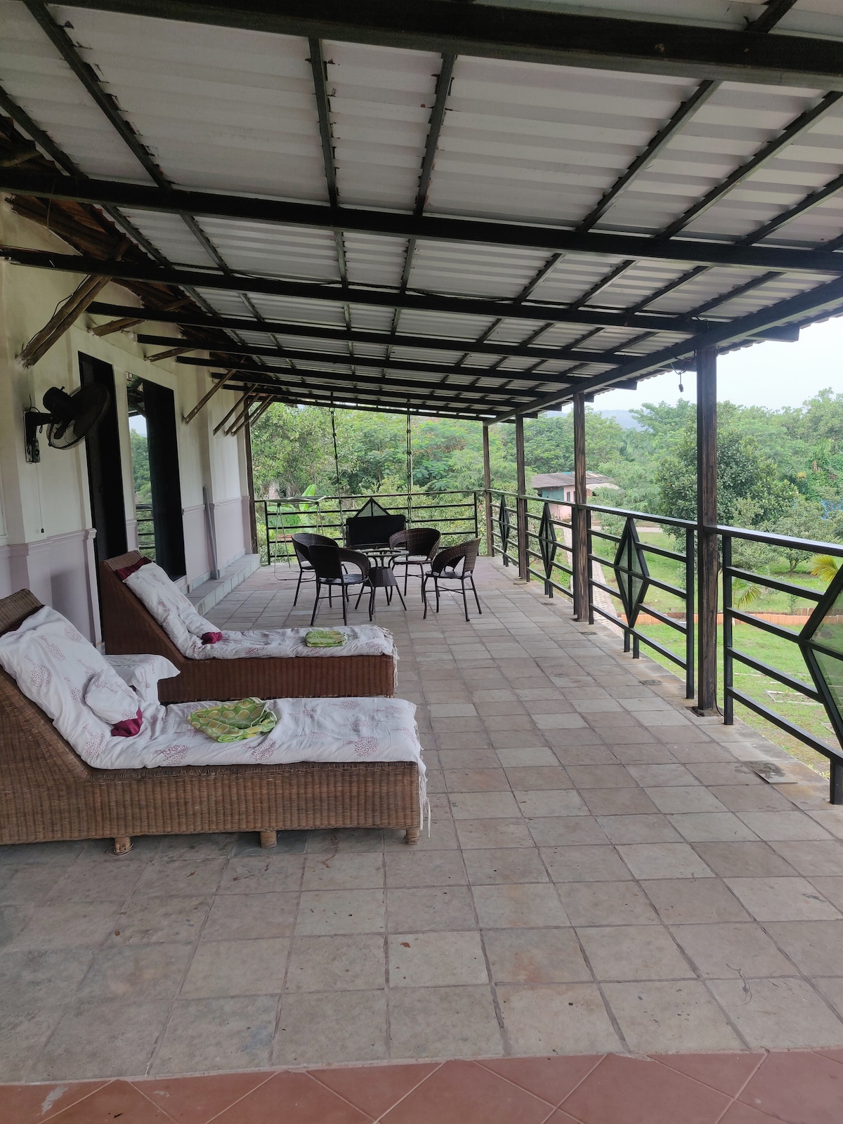 A spacious patio is displayed, featuring two lounge chairs and a dining table surrounded by chairs. The roof provides cover, while greenery is visible in the background through the railing. Light-colored tiles form the flooring, offering an open, airy feel.