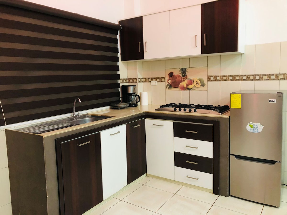 A modern kitchen is presented with wooden cabinetry featuring a combination of dark and light tones. A stainless steel refrigerator and a gas stove are visible, along with a coffee maker on the counter. Neutral tile flooring and striped window blinds complete the design.