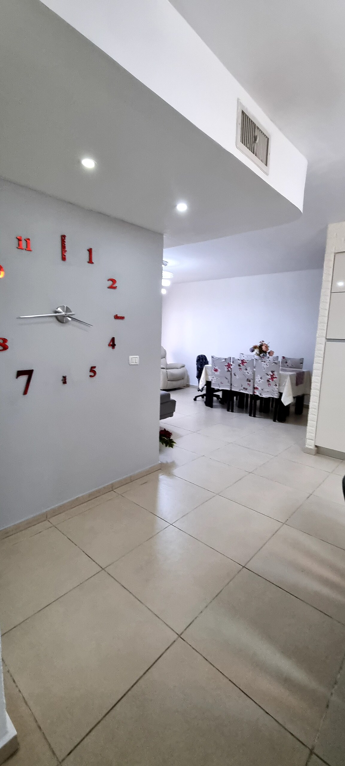 A hallway is depicted with a large wall clock displaying red numbers. Soft lighting illuminates the space, leading to an inviting room with a dining table set elegantly. A comfortable seating area is visible in the background, enhancing the open layout.
