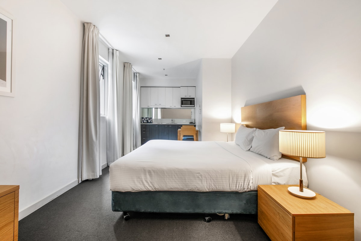 A bright and minimalist studio room features a spacious queen-sized bed flanked by stylish bedside tables, each with a lamp. An open kitchenette is visible in the background, accompanied by natural light filtering through the sheer curtains.