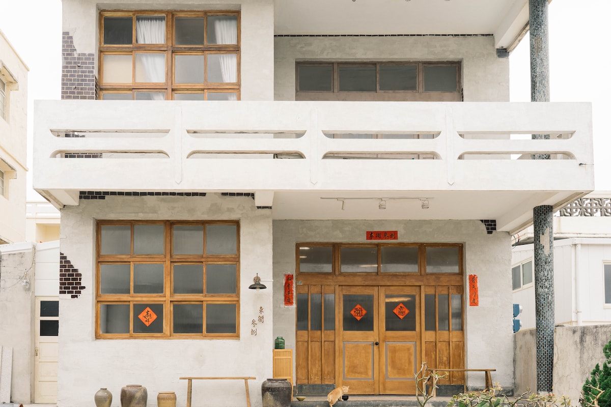 The exterior of the building features a modern design with large wooden windows and a spacious balcony. Traditional red decorations adorn the entrance, flanked by circular tables and minimal greenery, enhancing the welcoming presence of the property.