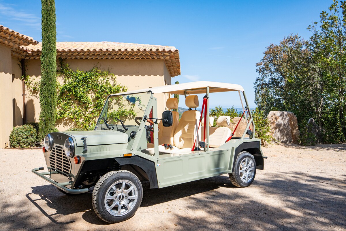 An electric Mini Moke is parked on a pebbled path, showcasing its light green exterior and cream seating. The backdrop features a villa with rustic charm, surrounded by lush greenery and a clear blue sky.