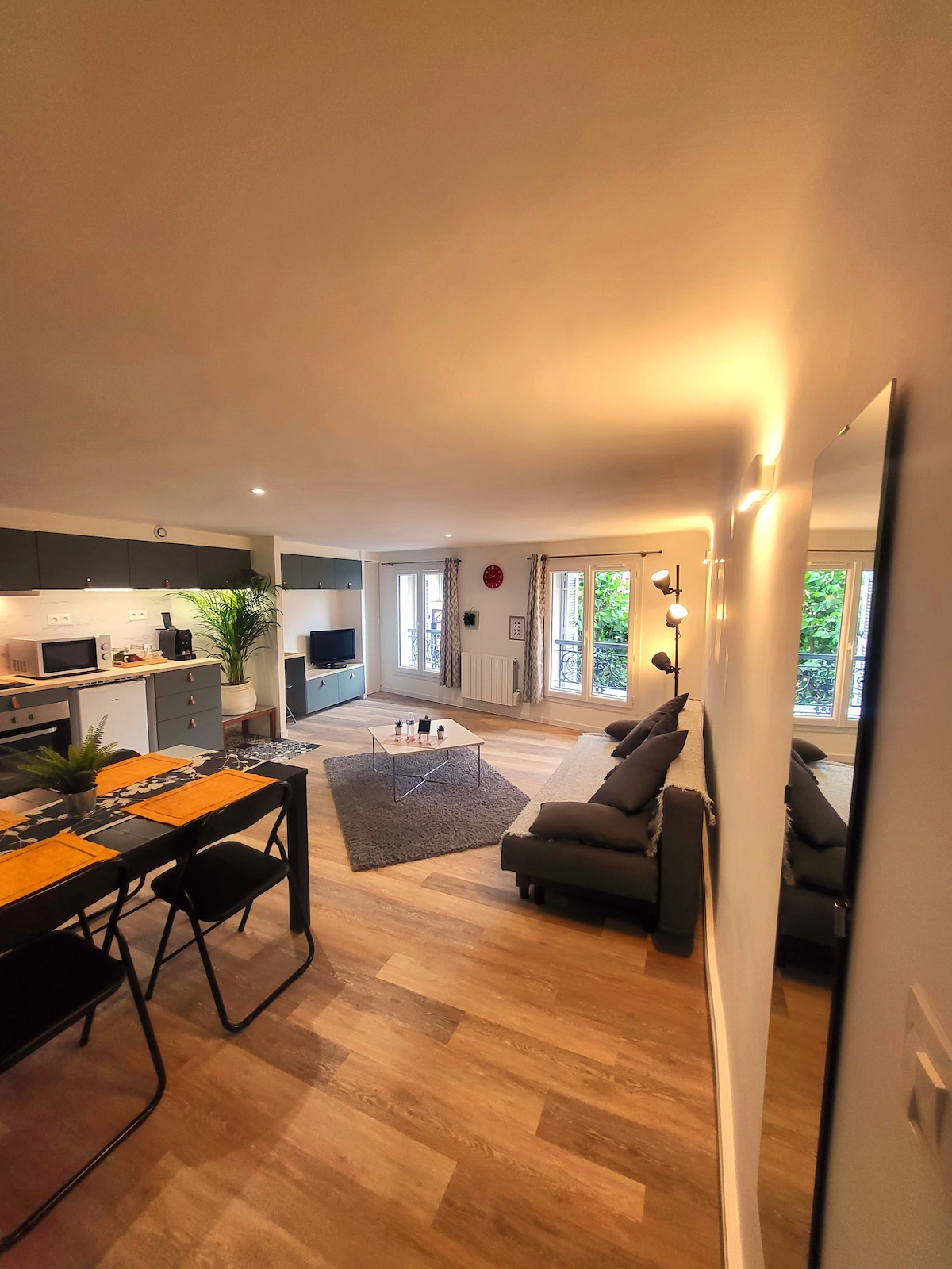The living area features an open layout with light-colored wood flooring. A modern gray sofa faces a coffee table, with a rug beneath. A dining table with black chairs stands nearby. Large windows allow natural light and offer views of greenery outside.