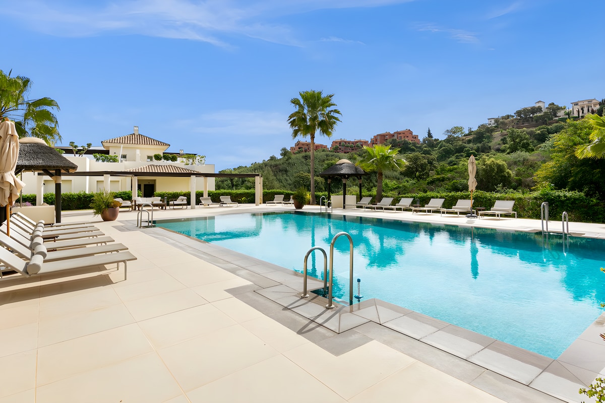 A spacious outdoor swimming pool is surrounded by lounge chairs and palm trees, offering a serene atmosphere. The pool features clear blue water, with steps leading into it. In the background, lush greenery and hillside properties enhance the tranquil setting.