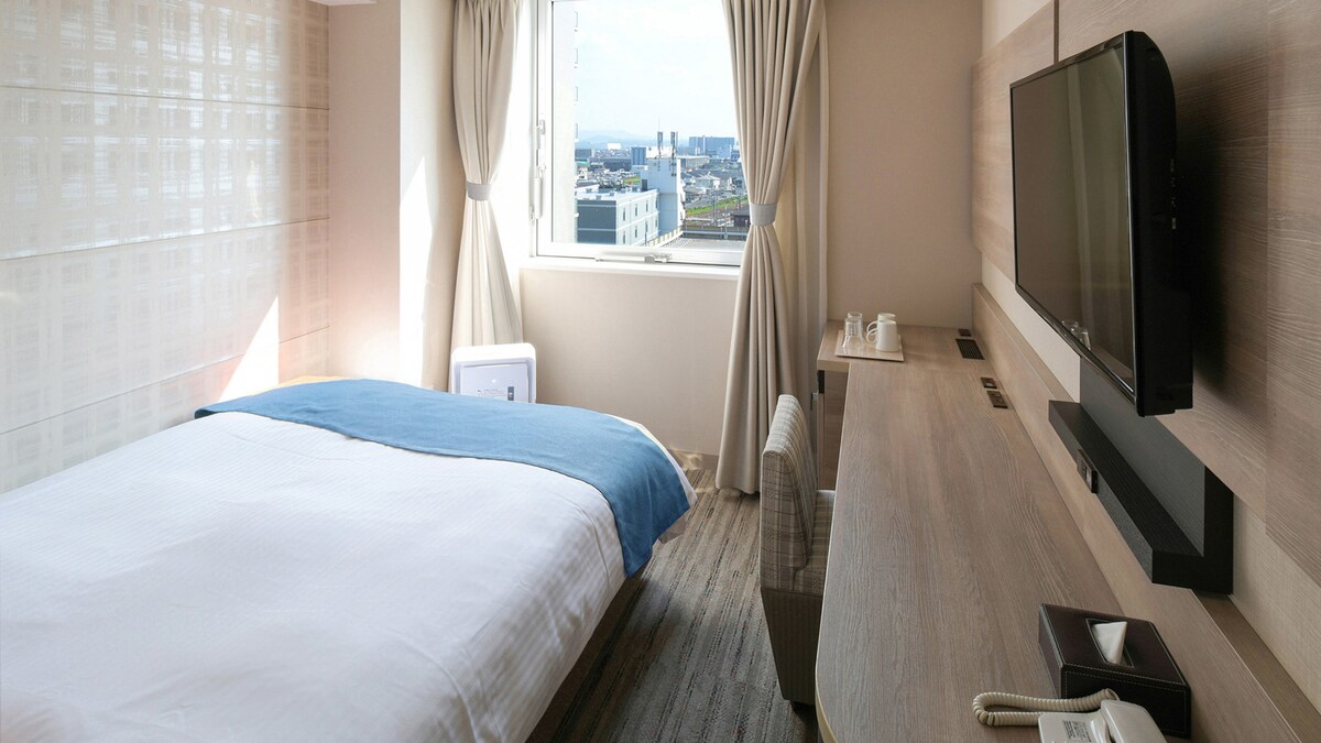 A standard single room features a semi-double bed with a crisp white duvet and a blue throw. A desk with a chair is positioned near a window, offering views of the surrounding landscape. Light filtering through the curtains enhances the room's modern decor.