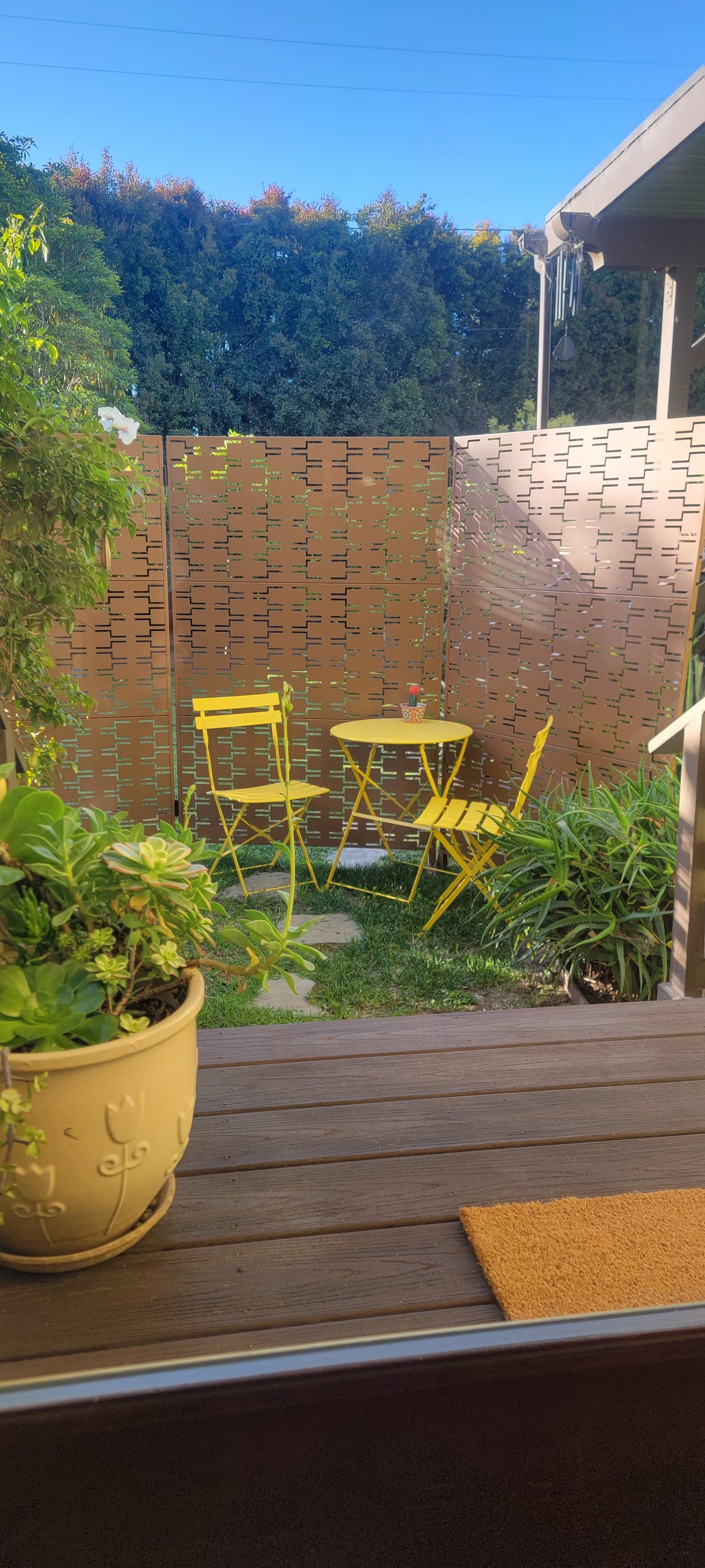 A private fenced patio features a wooden deck leading to a small grassy area. Bright yellow chairs and a matching table are surrounded by lush greenery and a decorative planter, providing a serene outdoor space for relaxation.